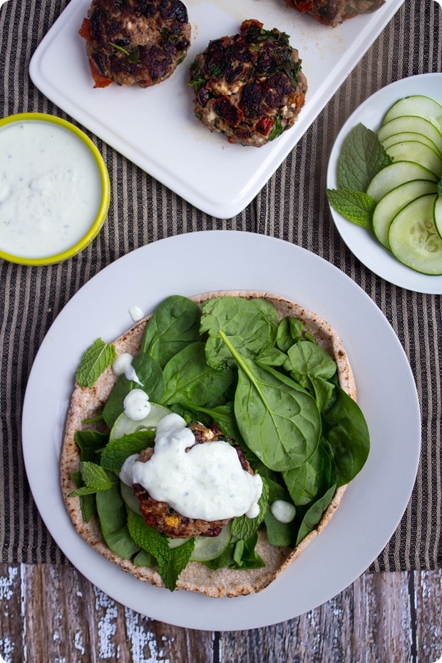 greek lamb burgers 6 white plates with cucumbers, spinach, and Greek lamb patties next to a small bowl of yogurt sauce