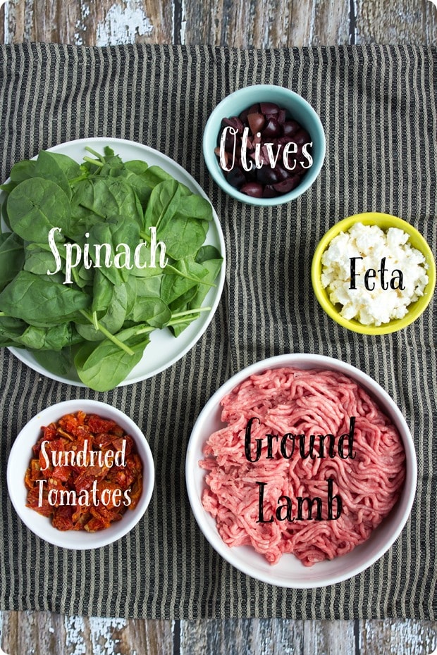 greek lamb burgers ingredients ground lamb, sun-dried tomatoes, feta, spinach, and olives in small bowls on a striped kitchen towel