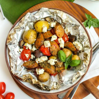 a foil packet with grilled potatoes, chicken sausage, vegetables, and crumbled feta cheese on a plate