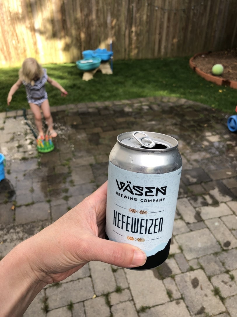 beer can with a toddler playing in a sprinkler in background