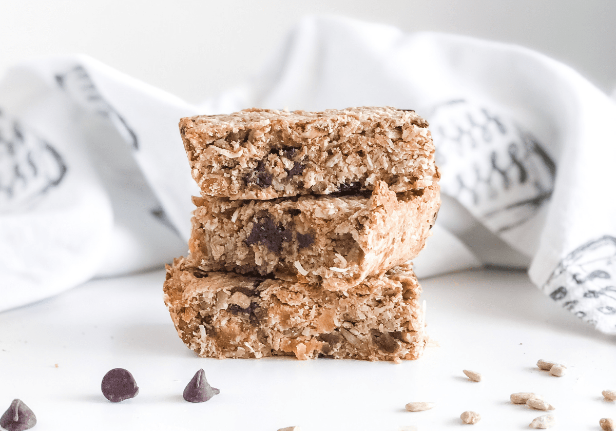 granola bars stacked with chocolate chips and sunflower seeds scattered in front and a kitchen towel behind