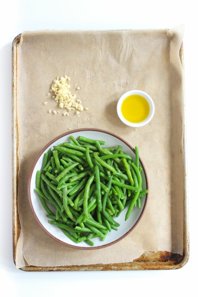frozen green beans with olive oil and garlic in a bowl on a sheet pan lined with parchment paper