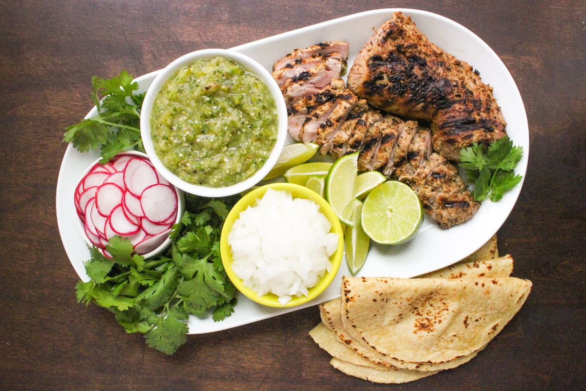 salsa verde in a bowl with other taco toppings, sliced pork, and tortillas