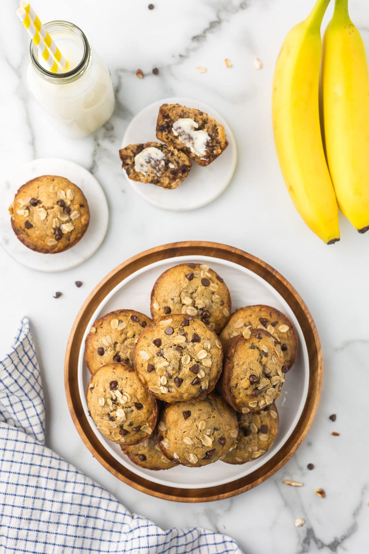 plate with a pile of gluten free banana muffins next to a glass of milk and a kitchen towel on a marble surface