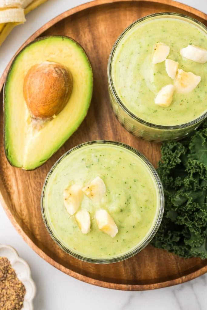 two green smoothies in short glasses with sliced banana on top sitting on a wooden platter