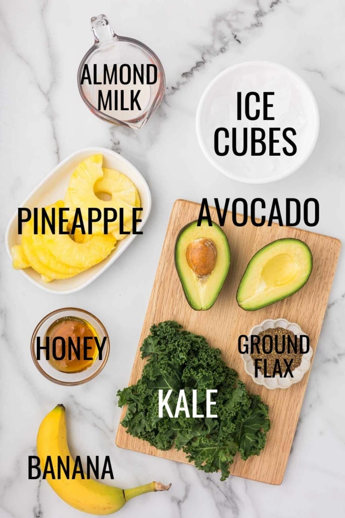 avocado, kale, pineapple, banana, ground flax, almond milk, and ice cubes in small bowls on a marble countertop